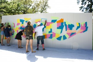 Graffiti artist and founder of Aerosol Warfare, GONZO247 leads students in spray painting a blank white wall propped up against the fence in the plaza.